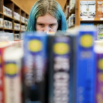 A person with blue hair seen beyond a shelf of books.