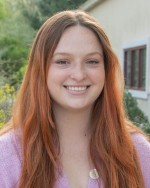 Headshot of Hannah Mennella. Hannah is an adult female with long auburn hair and is wearing a pink sweater.