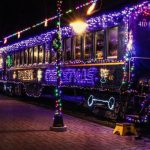 A festive train adorned with colorful Christmas lights, glowing brightly against the night sky. The train features intricate decorations, including wreaths and illuminated patterns, creating a cheerful holiday atmosphere. A brick-paved platform is visible in the foreground.